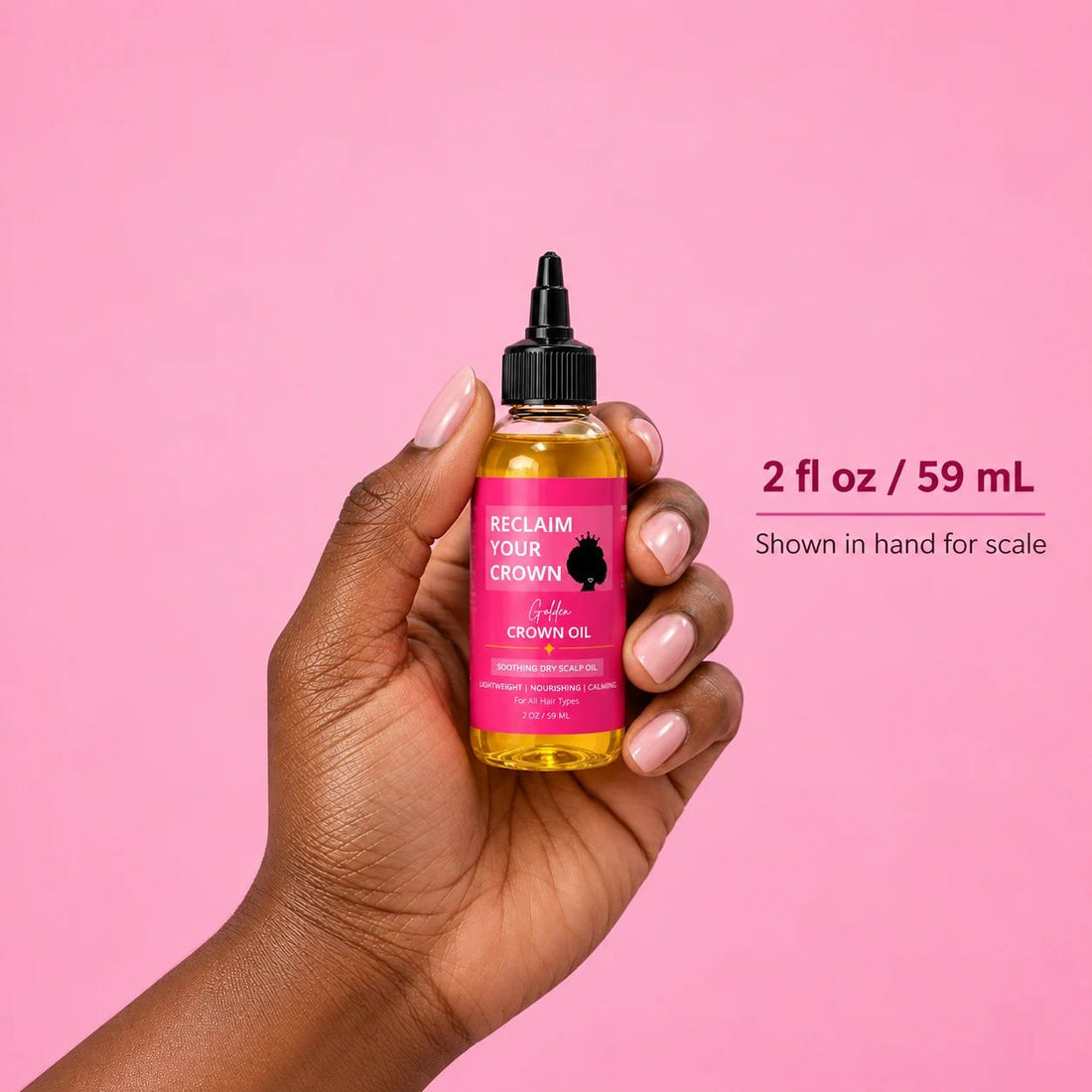 A hand holding a bottle of Reclaim Your Crown Oil against a solid pink background, with text indicating the volume is 2 fl oz / 59 mL.