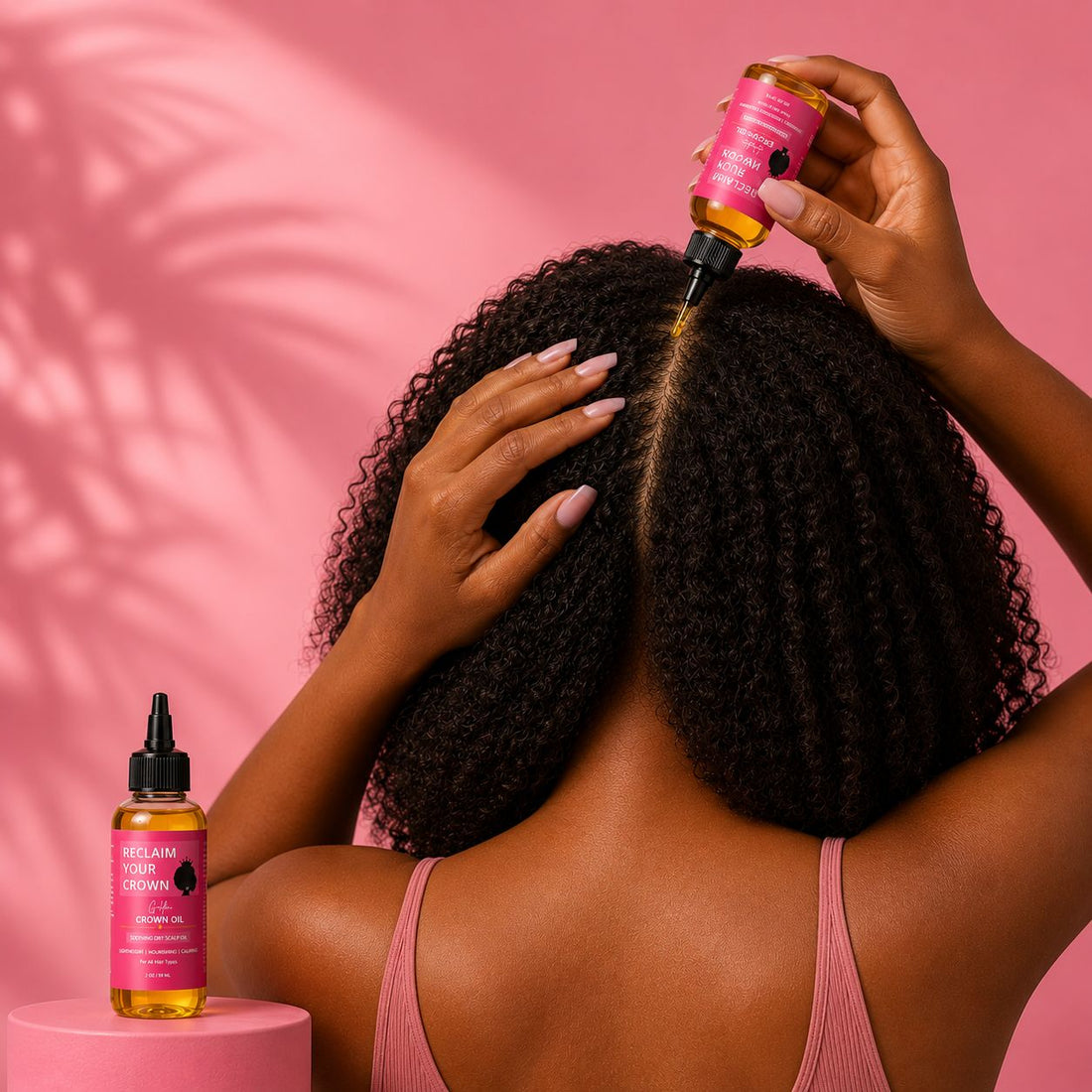 Black woman with curly hair applying Golden Crown Oil directly to her scalp against a soft pink background, with a 2 oz Reclaim Your Crown hair oil bottle displayed on a pink pedestal.