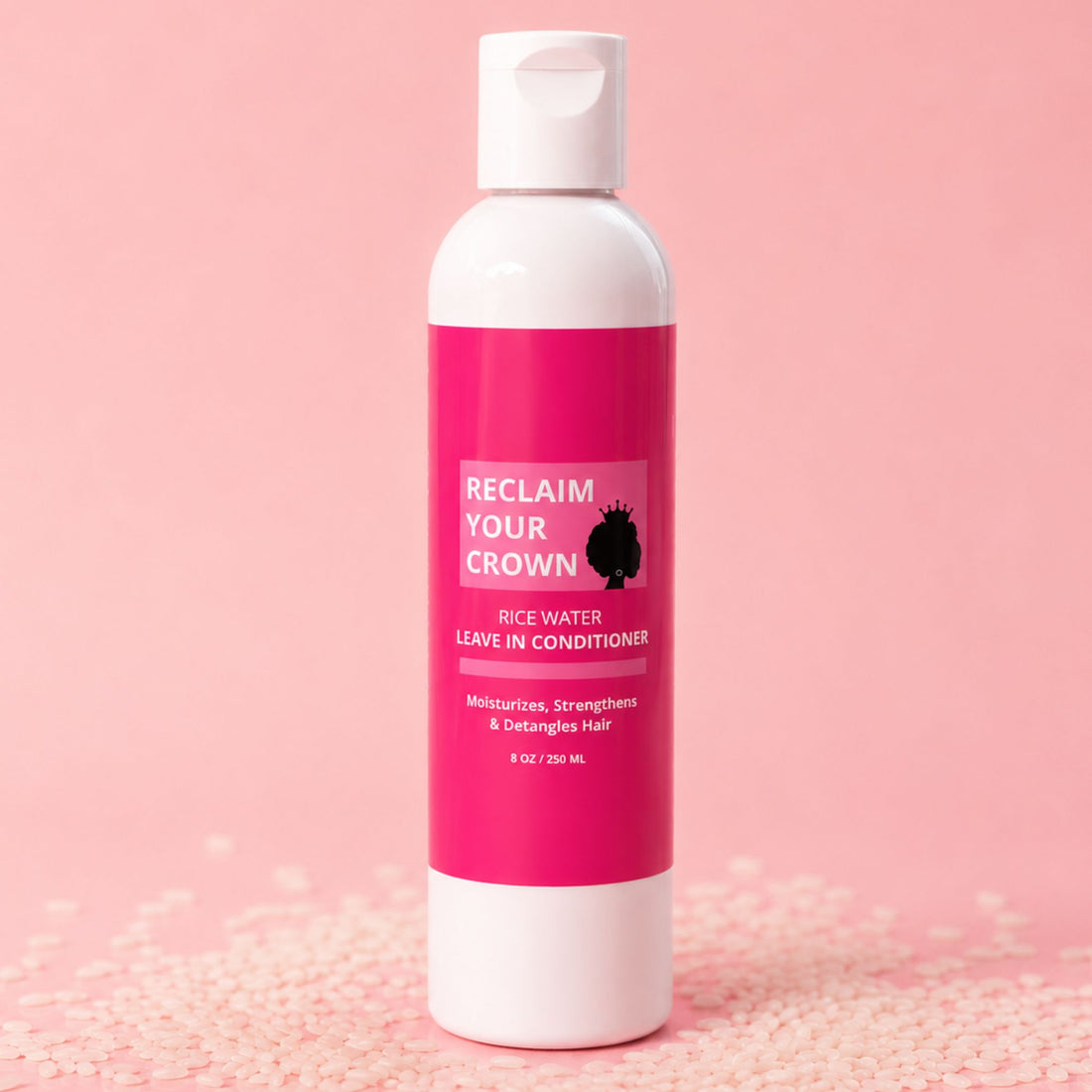 Bottle of 'Reclaim Your Crown' rice water leave-in conditioner on a pink background surrounded by rice.